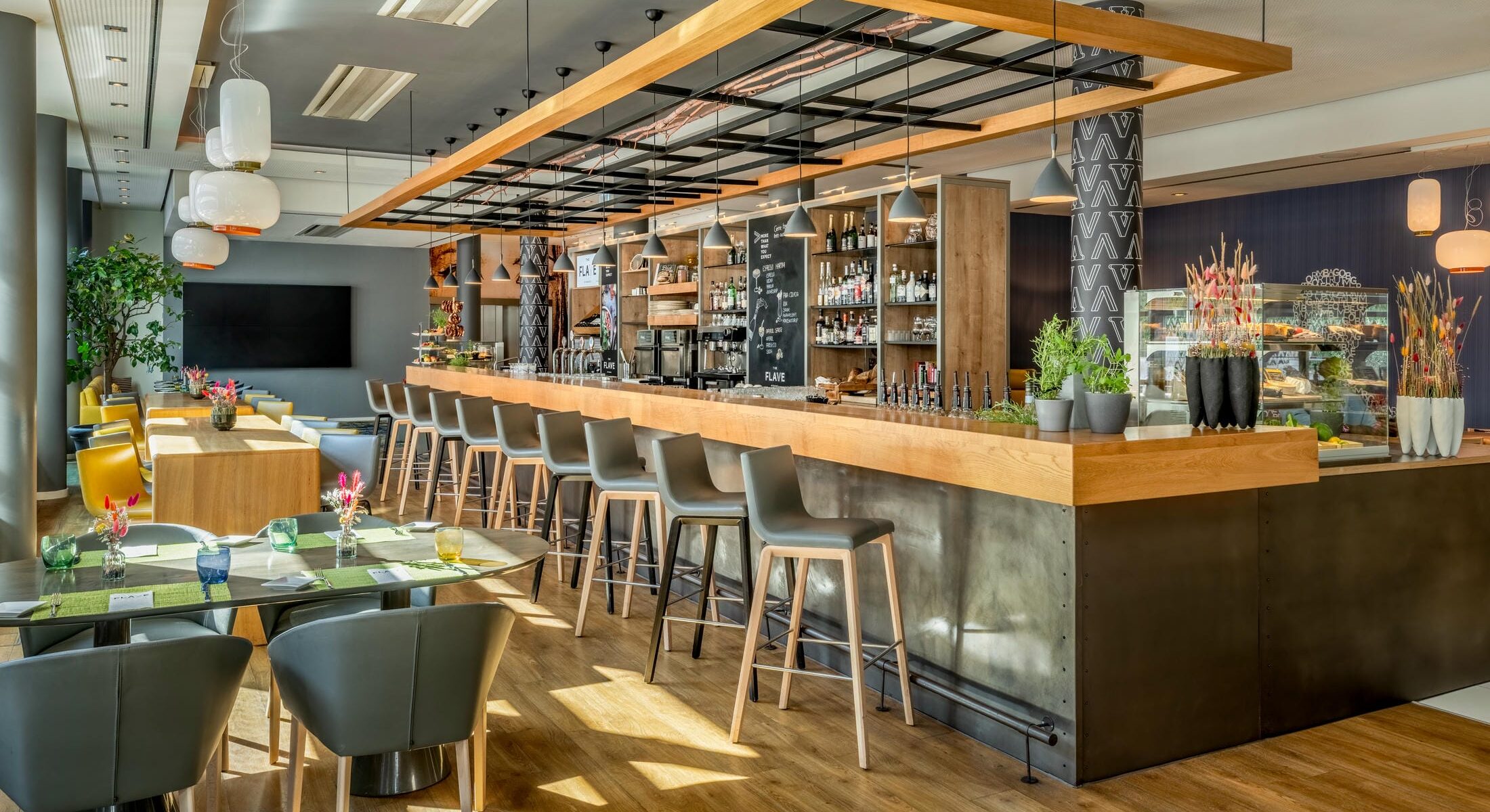 Barbereich des The Flave of Munich. im Novotel München City mit warmem Holzboden, großer TV-Wall, Hochtischen und Bar.