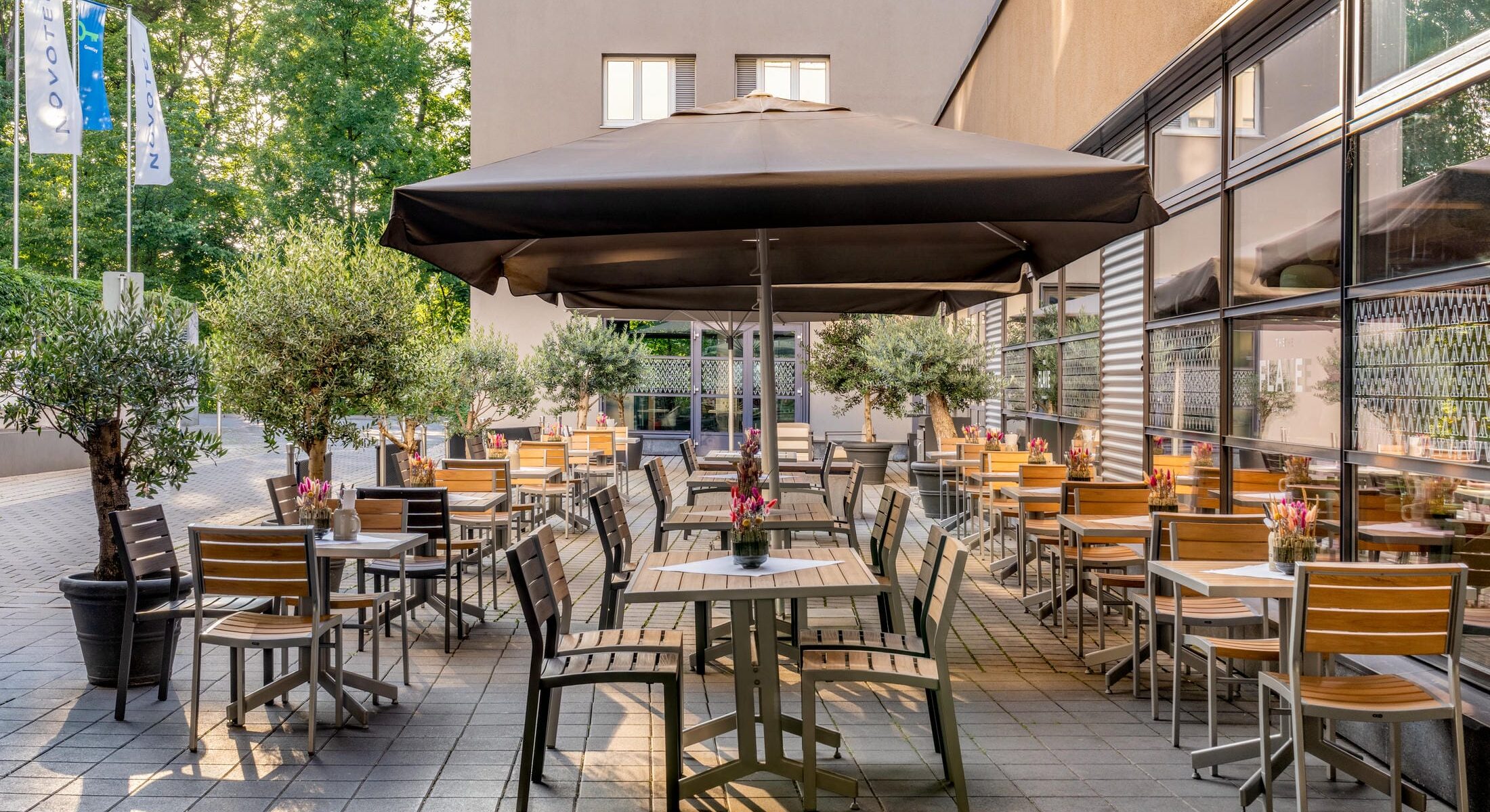 Sonnendurchflutete Terrasse mit Restaurantbestuhlung und Sonnenschirmen und vielen Bäumen rundherum im Novotel München City