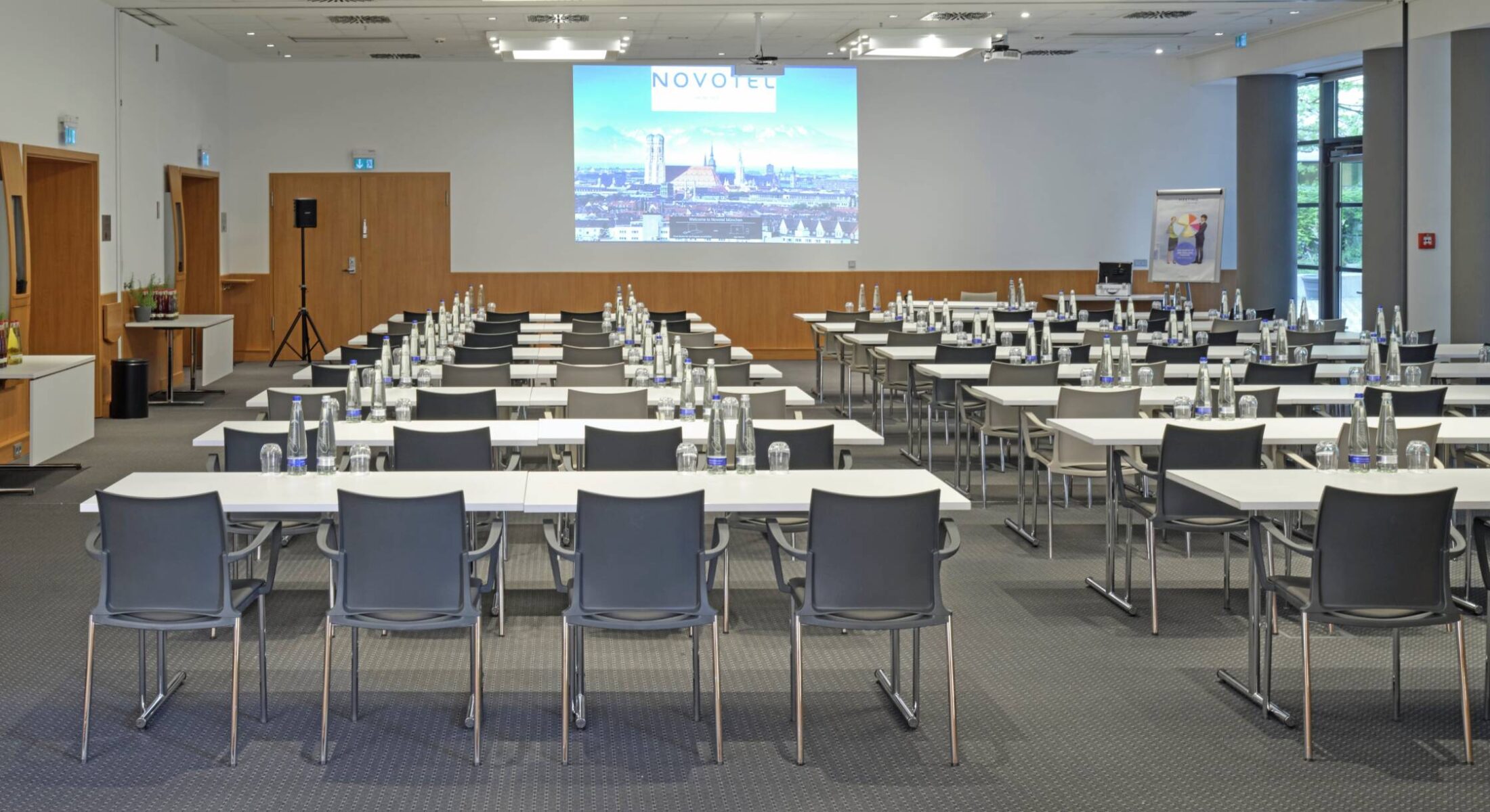 Tagungsraum mit parlamentarischer Bestuhlung und großer Leinwand zur Projektion sowie viel Tageslicht im Novotel München City